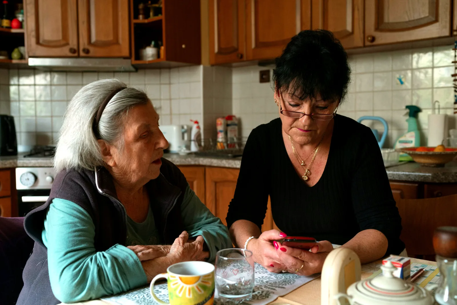 An adult daughter sits beside her mother at a warm kitchen table, showing her something on a phone